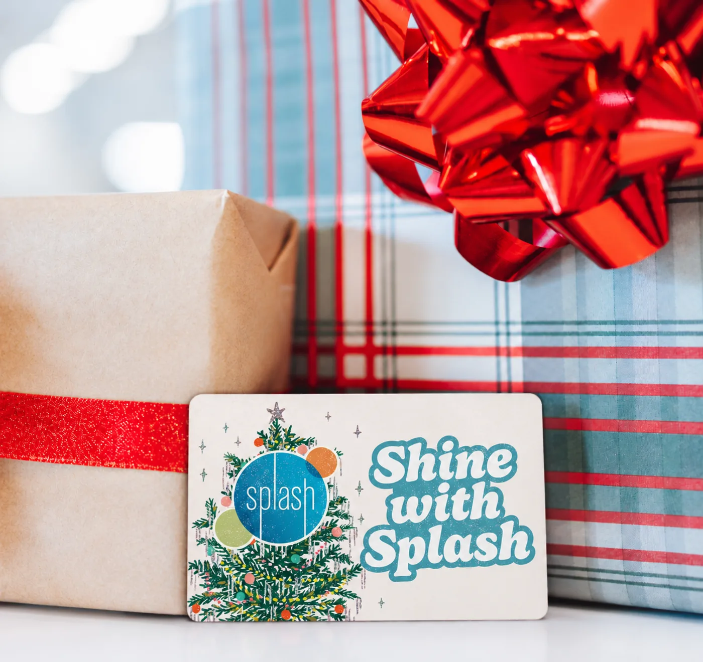 A Shine with Splash holiday gift card, decorated with a Christmas tree and the Splash logo, leaning against wrapped presents with a red bow