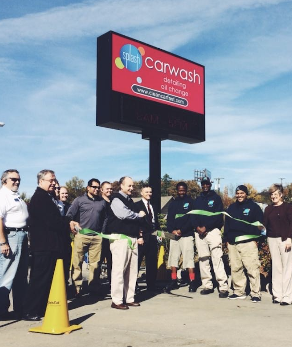 Splash Car Wash employees stand in a group for a ribbon cutting ceremony at a location in 2014. Above them is a red sign with the Splash logo.