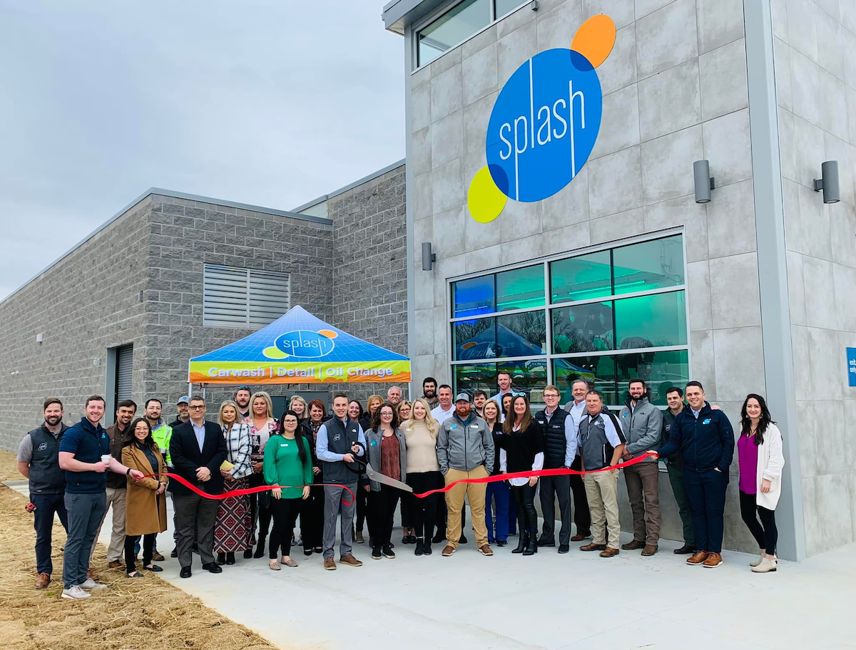 Group of Splash Employees stand in front of Splash Car Wash building for a ribbon cutting ceremony. The man at the front of the crowd hold giant scissors and has just cut the red ribbon, which is being held at each end by other employees at the edge of the group.
