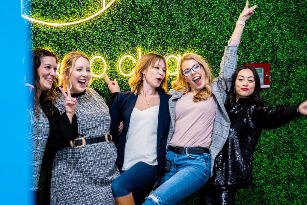 Five women pose together smiling in front of a greenery wall with a neon sign.