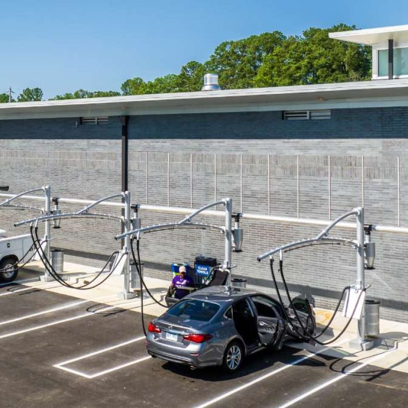 Four vehicles are parked at the outside Splash Car Wash vacuuming station, with two people standing outside of their vehicles and currently vacuuming. The vacuuming stations stand in front of a gray wall and have designated parking spots.