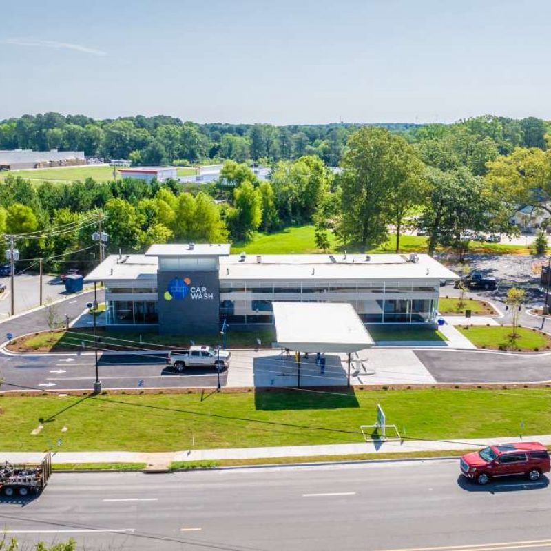 Outside view of front of Bryant Splash Car Wash gray and white building with Splash Car Wash logo on side. Parking lot of the building connects to the highway where vehicles are driving.