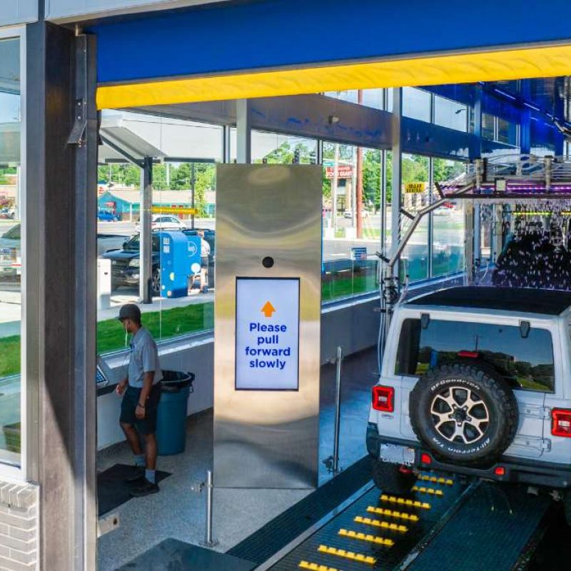 Water and soap are put on the white Jeep driving through the Bryant Splash Car Wash. A male Splash employee stands by the window looking at a tablet while the directions next to the entrance read "Please pull forward slowly."