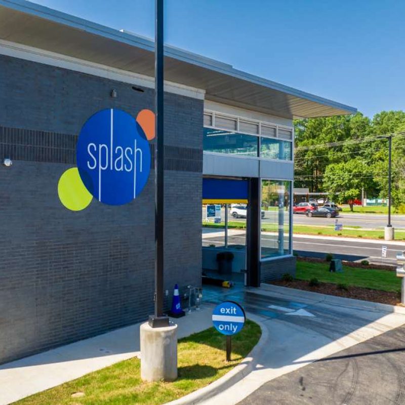 Blue circular sign sits outside of gray Splash Car Wash building and reads "exit only." An white arrow on the ground shows vehicles where to exit as they come out of the car wash.
