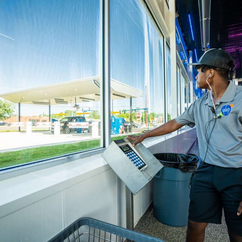 Male Splash Car Wash employee wears gray shirt with company logo and black shorts while standing inside facility. He looks out of window to the vehicles in line for the car wash.