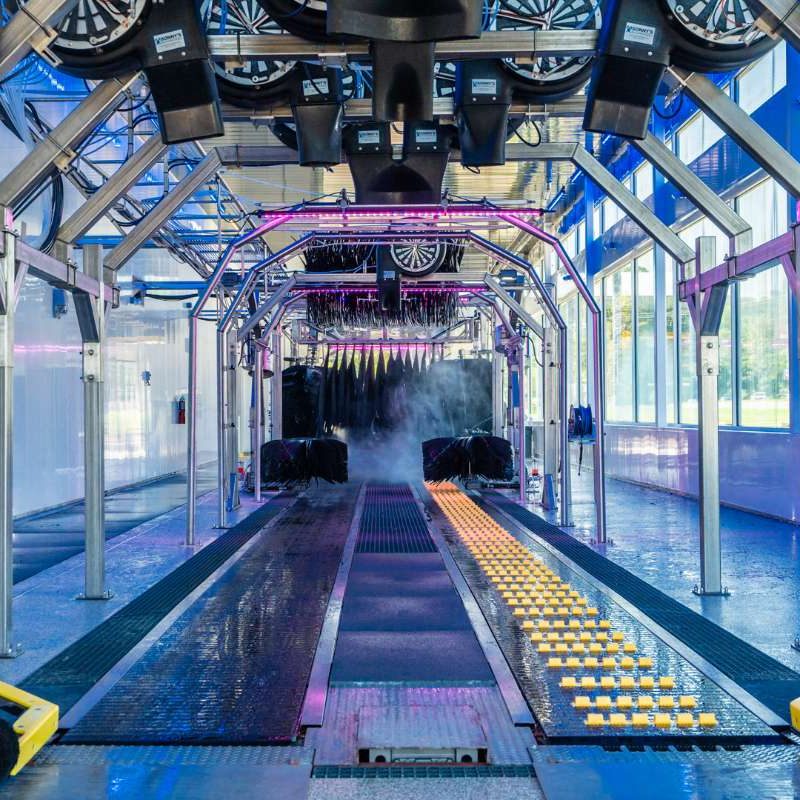 Inside of drying station at Bryant Splash Car Wash. Rollers/tracks are on the floor for cars to ride on through the car wash process. The room is lit up by natural light from windows and pink and blue lights inside.