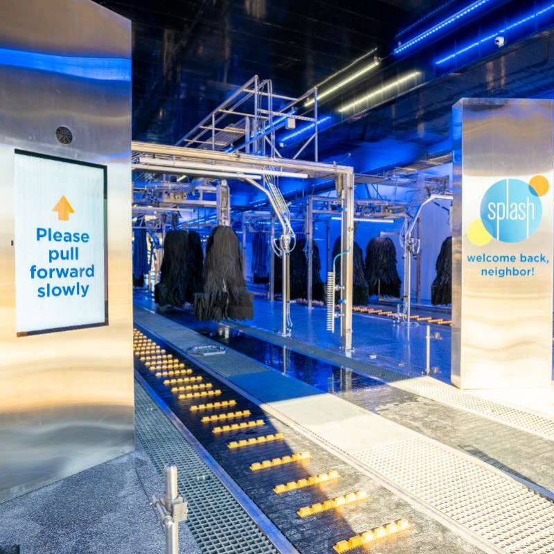 Entrance to the Splash Car Wash with rollers/tracks for vehicles to ride up on. Sign below the Splash logo reads "welcome back, neighbor!" while instructions on the other side of the tracks say "Please pull forward slowly."