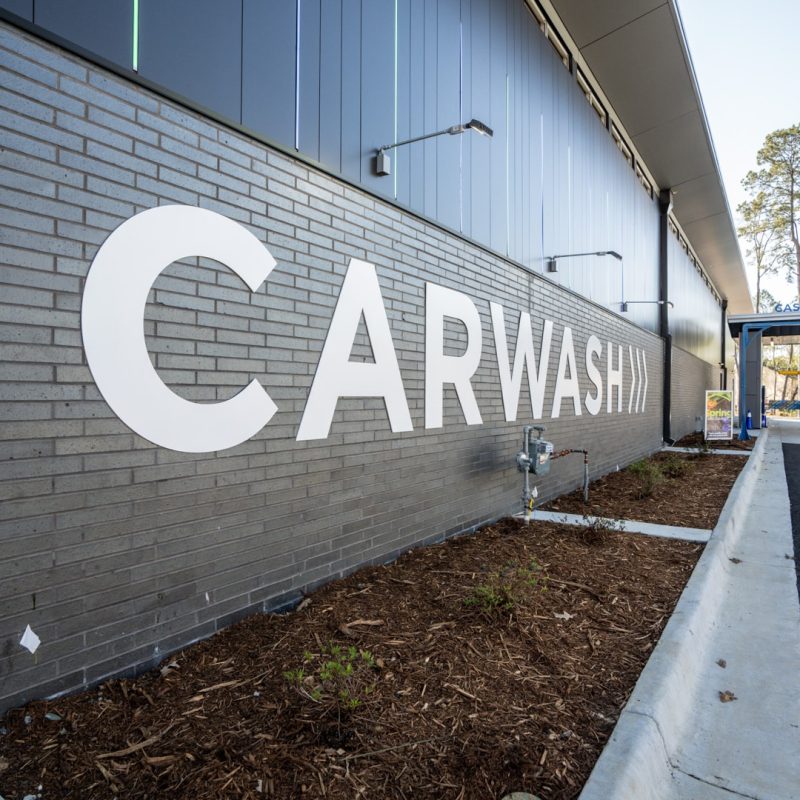 Side of Splash Car Wash building next to the entrance lanes is gray brick and siding with "CARWASH" written in big white letters with three arrows.