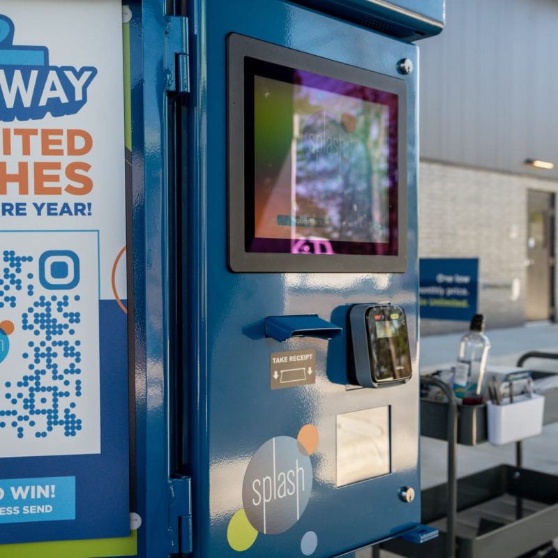 Blue Splash Car Wash payment kiosk outside of building. Sign on kiosk says "Giveaway: Unlimited Washes for an Entire Year! Enter to Win! Just Scan & Press Send" with a QR code in between the text.