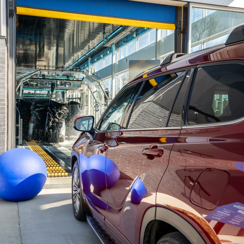 A red SUV waits to drive onto the conveyer belt and be pushed through the Sherwood Splash Car Wash. The brushes clean a car on the conveyer belt in the background.