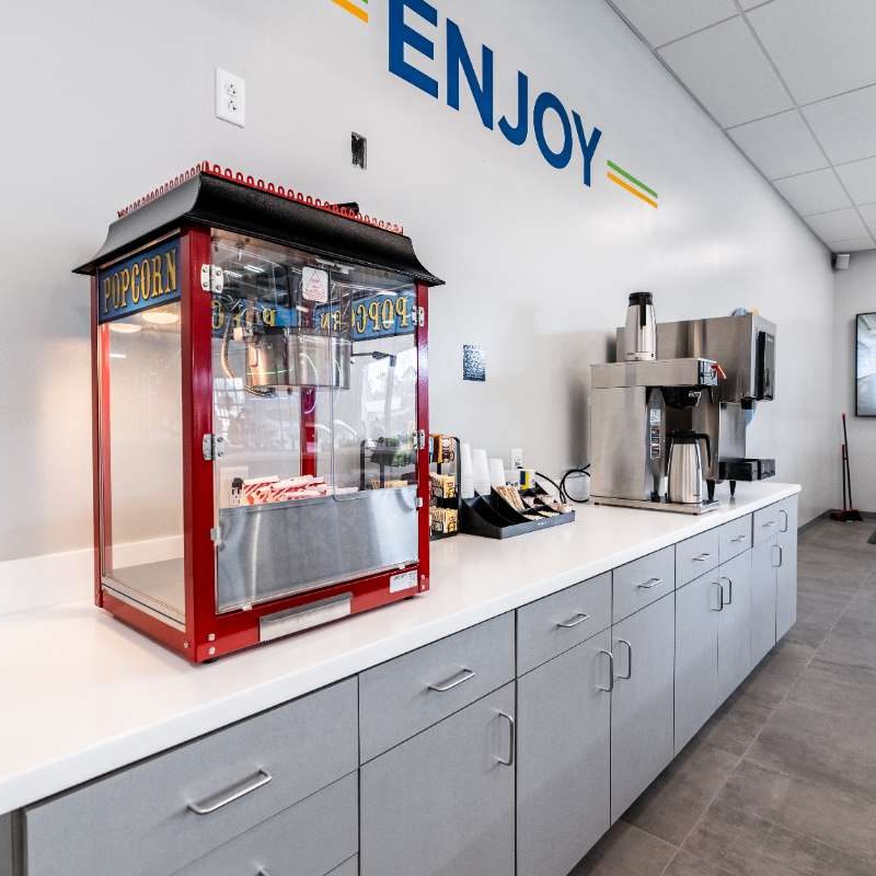Coffee/snack bar inside Splash Car Wash facility. "ENJOY" is written in blue on the white walls above a white countertop with gray cabinets. A drink fountain, coffee machine, and popcorn machine sit on the counter.