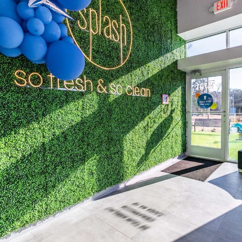 Inside lobby of Splash Car Wash. Small black couch is sitting in front of see-through wall and door facing the playground area outside. Adjacent wall is covered in greenery, blue balloons, the Splash logo, and "so fresh & so clean" is gold lettering.