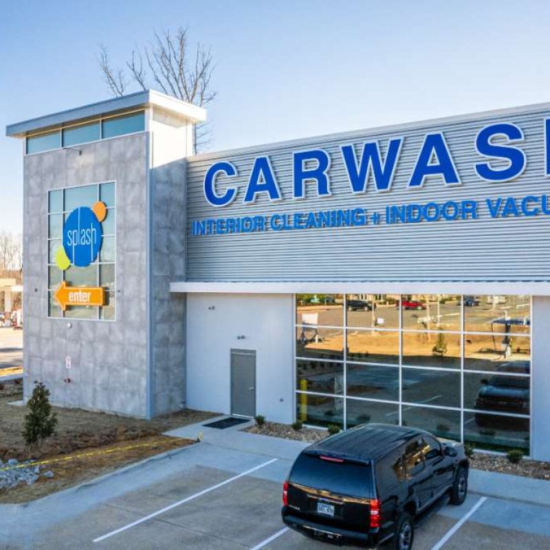 Outside of Splash Car Wash West Little Rock facility. White and gray building with blue text reading "CARWASH INTERIOR CLEANING + INDOOR VACUUMS." Two vehicles are parked next to building.